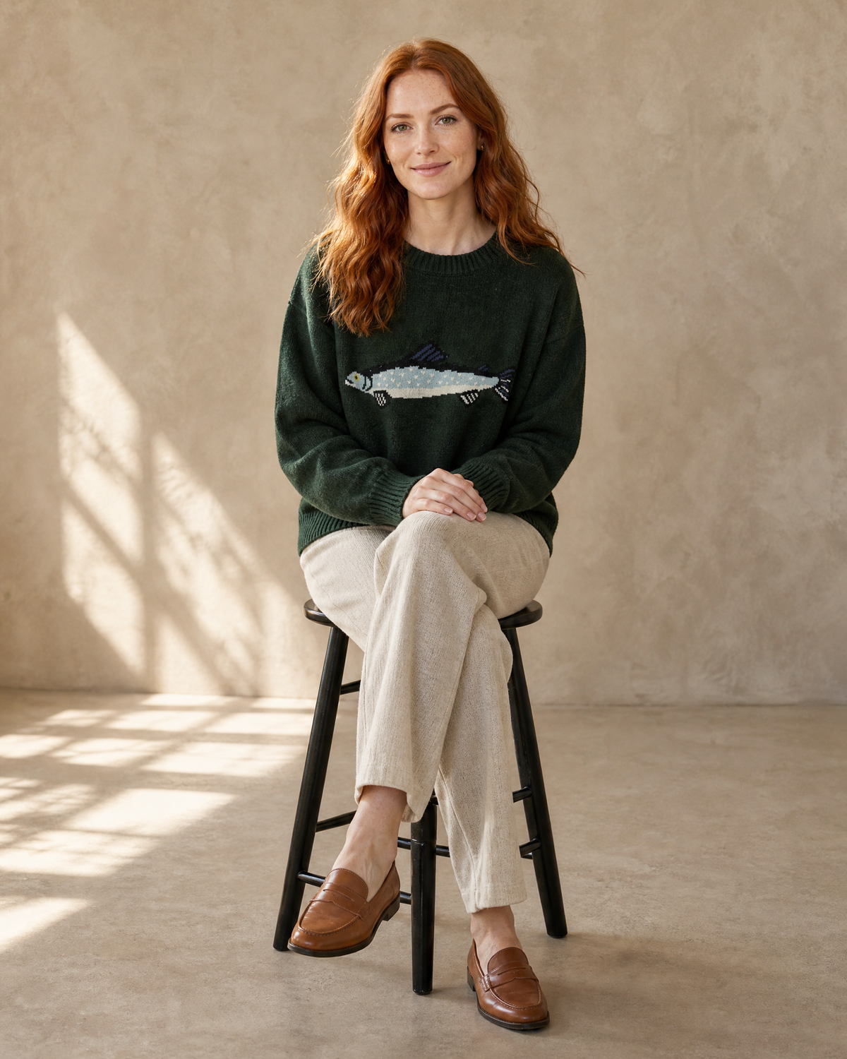 Women's Relaxed Fish Motif Knit Sweater in Dark Green, stool, legs crossed, worn by model